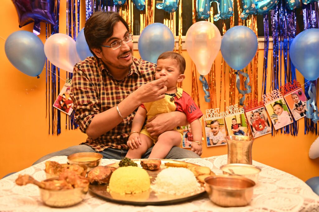 pexels-photo-19962115-19962115 A joyful father feeds his baby son during a vibrant Indian birthday celebration with traditional food and decorations.